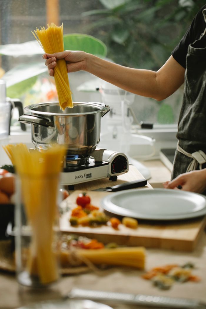 Services Crop unrecognizable female chef in apron putting raw spaghetti into saucepan on stove while cooking in contemporary light kitchen