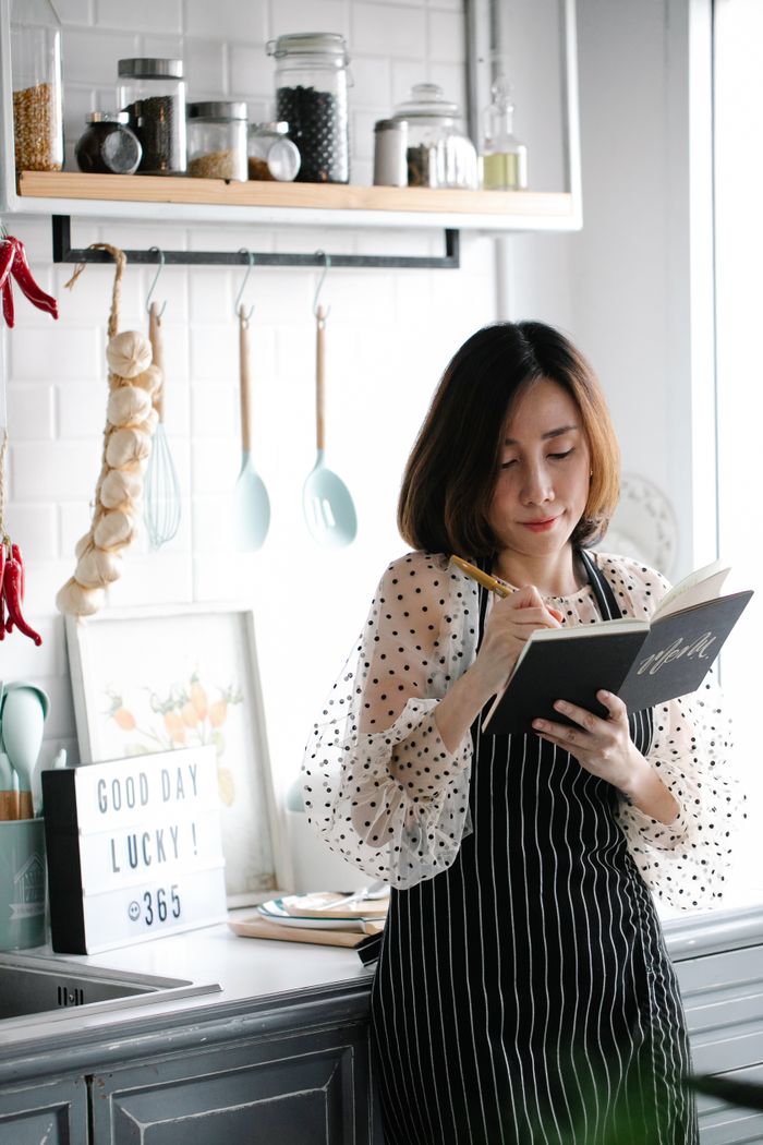 Services Asian woman in stylish apron writing in a cookbook in a modern kitchen setting.